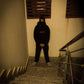 Person wearing a dark hoodie and pants standing on a dimly lit staircase.