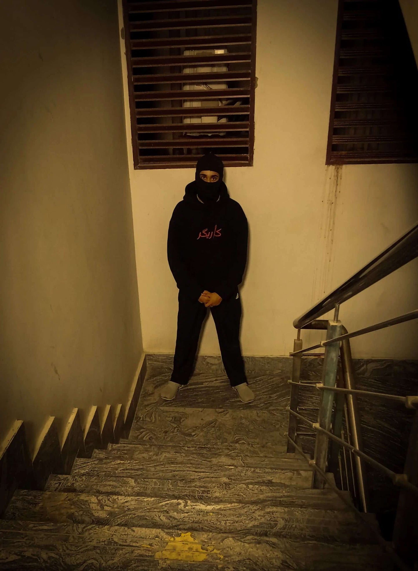 Person wearing a dark hoodie and pants standing on a dimly lit staircase.