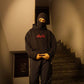 Person wearing a black hoodie with red text and balaclava standing on a staircase.
