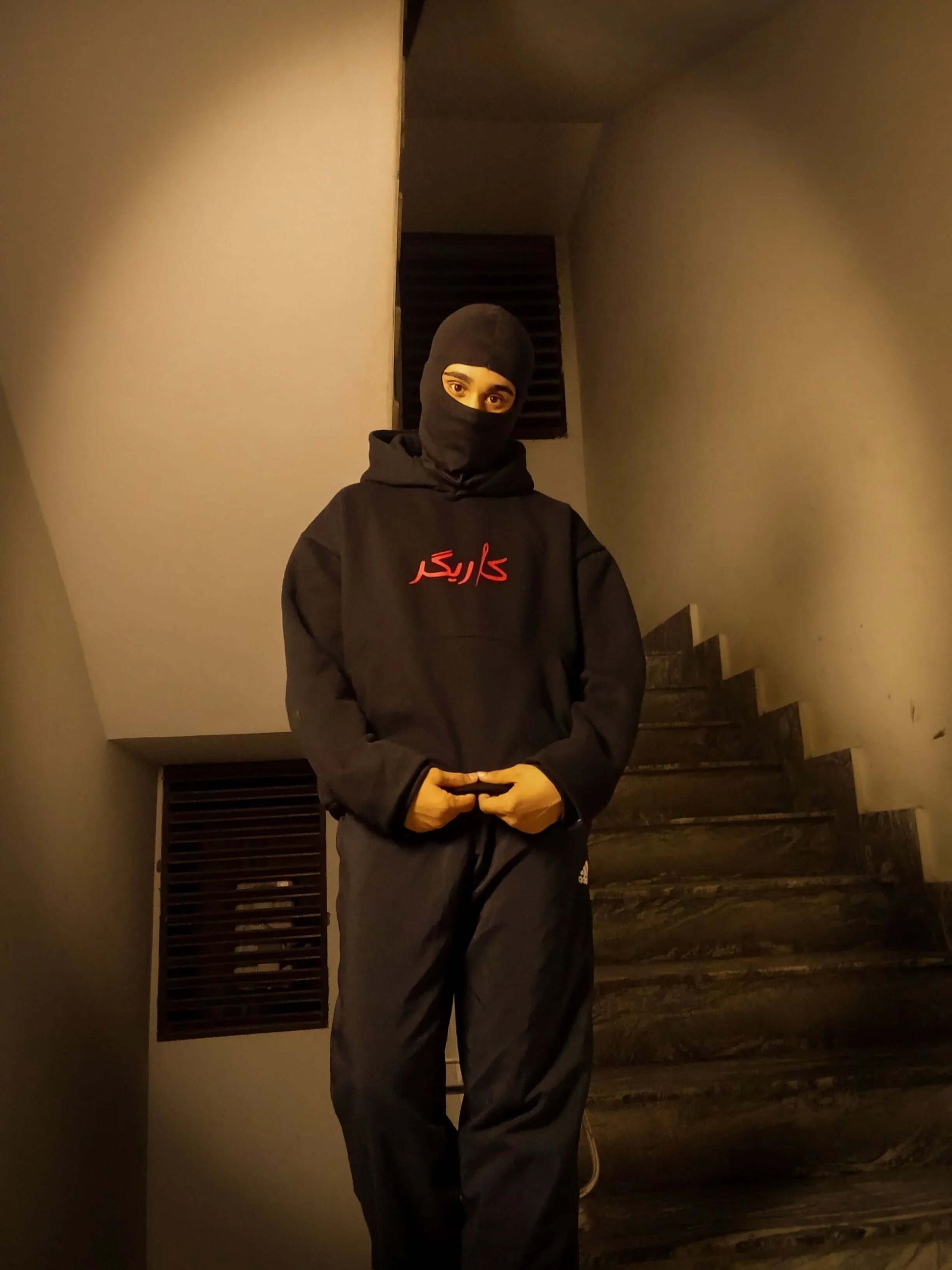 Person wearing a black hoodie with red text and balaclava standing on a staircase.